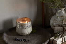 The nkuku Juniper & Fir 3 Wick Scented Candle, in a rustic textured holder, sits on a wooden tray near dried berries and gifts. A ceramic vase with greenery adds to the cozy, natural scene and soothing winter scent.