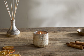The nkuku Mandarin & Clove 1 Wick Scented Candle rests in a ceramic holder on a rustic wooden table, surrounded by dried orange slices, cinnamon sticks, and a vase with reed diffusers against a neutral wall.