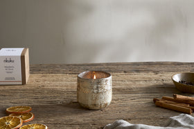 A nkuku Mandarin & Clove 1 Wick Scented Candle in a rustic holder sits on a wooden table with dried orange slices, cinnamon sticks, and cloves, filling the air with its festive fragrance.