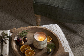 The nkuku Mandarin & Clove 1 Wick Scented Candle in a rustic holder sits on a wooden tray with dried orange slices and greenery, filling the air with festive fragrance beside a green chair on a textured rug.