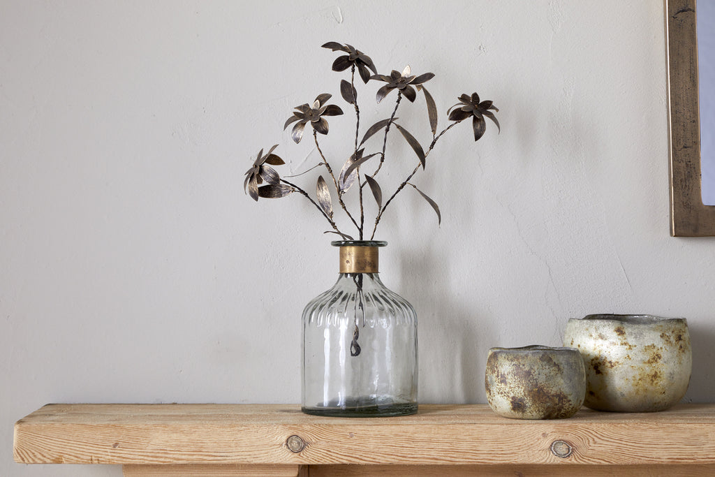 The Svara Stem Flower - Antique Brass by nkuku adds elegance to a clear glass vase. Nearby, two small rustic ceramic pots rest on a wooden shelf against a plain, light-colored wall.