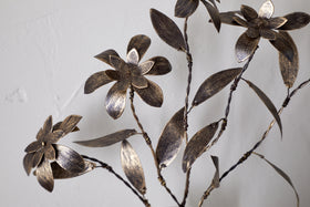 Close-up of the nkuku Svara Stem Flower, a metallic sculpture with stylized floral and leaf motifs, featuring an antique brass stem, set against a plain white background.