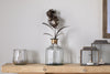 The Svara Stem Hyacinth in Antique Brass by nkuku stands in a glass vase on a wooden shelf beside a small glass tumbler and two metal lanterns with lit candles, set against a plain white wall.