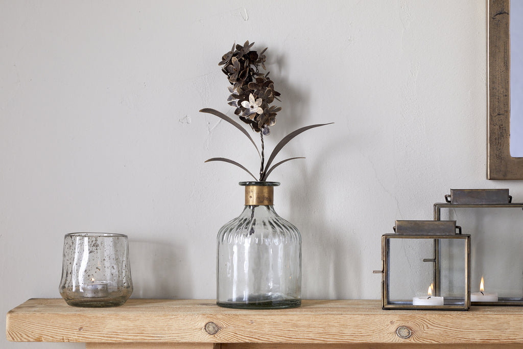 The Svara Stem Hyacinth in Antique Brass by nkuku stands in a glass vase on a wooden shelf beside a small glass tumbler and two metal lanterns with lit candles, set against a plain white wall.