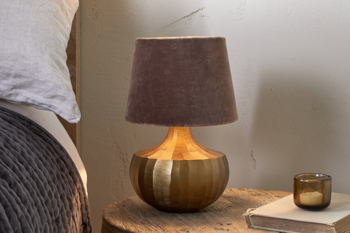 A bedside table displays the nkuku Sakiti Cast Aluminium Table Lamp in antique brass with a soft brown velvet shade, beside a small amber glass candle holder and old book. The gray quilted edge of a bed is visible on the left.