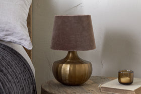 The Sakiti Cast Aluminium Table Lamp in Antique Brass by nkuku, featuring a textured round base and soft brown velvet shade, sits on a rustic nightstand beside gray bedding, with a closed book and brown glass candle holder nearby.