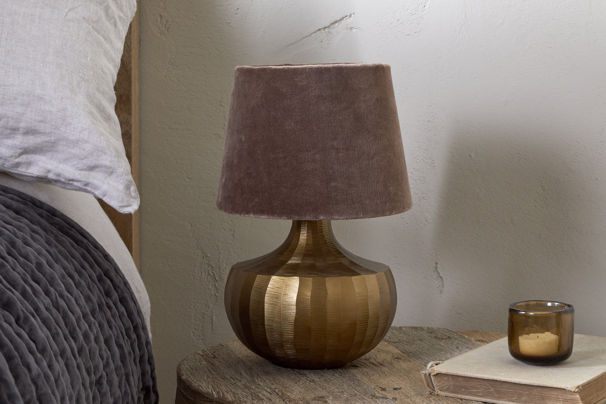 The Sakiti Cast Aluminium Table Lamp in Antique Brass by nkuku, featuring a textured round base and soft brown velvet shade, sits on a rustic nightstand beside gray bedding, with a closed book and brown glass candle holder nearby.