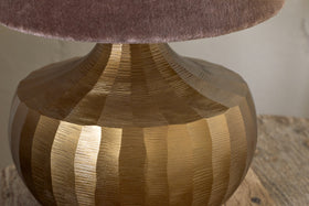 Close-up of the Sakiti Cast Aluminium Table Lamp by nkuku in antique brass, featuring a textured base with vertical ridges and topped with a soft, plush mauve lampshade on a rustic wooden surface.