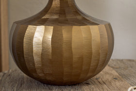 A close-up of the nkuku Sakiti Cast Aluminium Table Lamp in antique brass, featuring a round shape and alternating matte and shiny gold vertical stripes, sits on a textured wooden surface against a softly blurred neutral background.