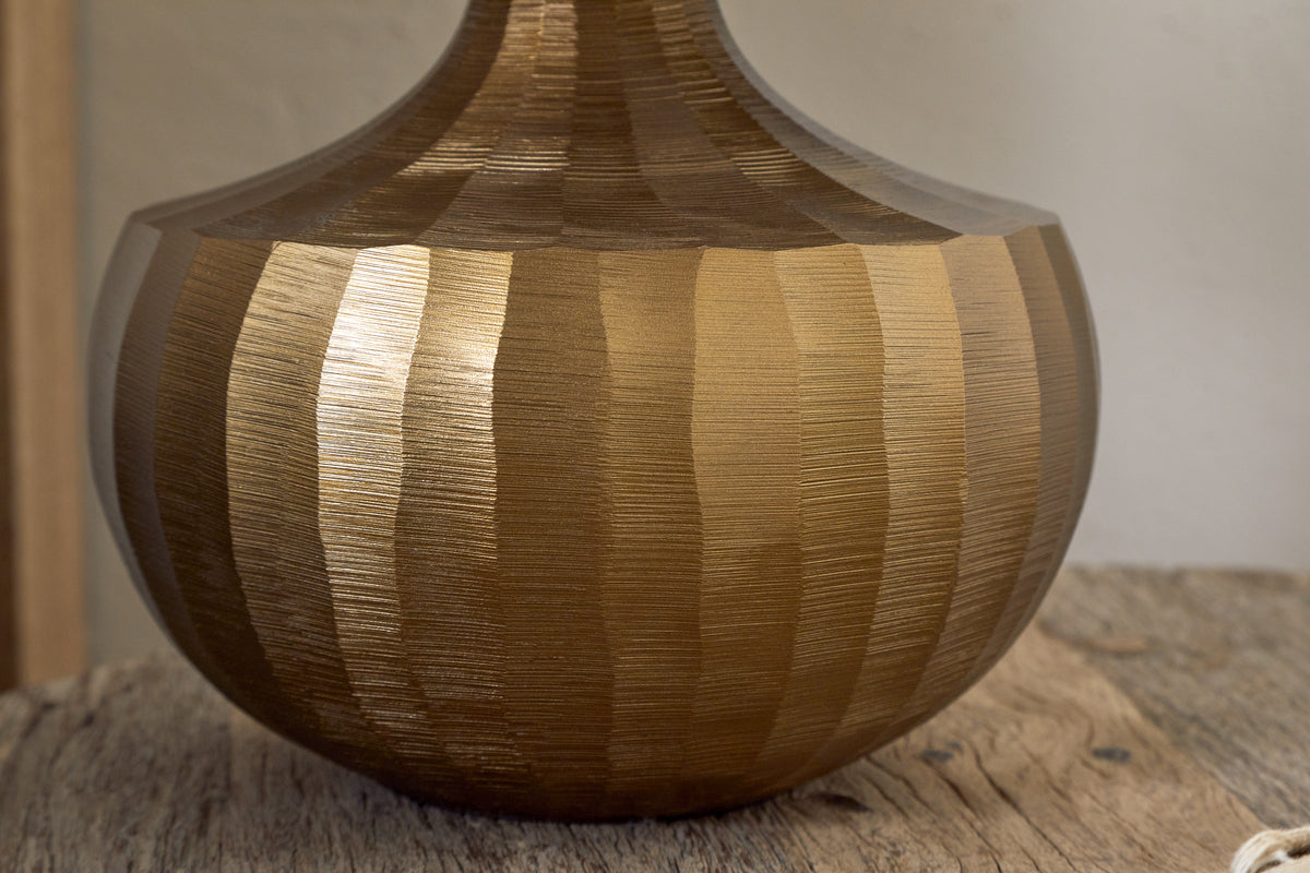 A close-up of the nkuku Sakiti Cast Aluminium Table Lamp in antique brass, featuring a round shape and alternating matte and shiny gold vertical stripes, sits on a textured wooden surface against a softly blurred neutral background.
