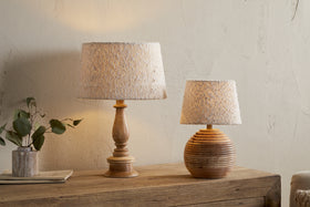 Two Sakiti Bouclé Lampshade – Off White by nkuku lamps add ambient light atop a rustic dresser. One lamp has a tall carved wooden base, the other a round ribbed base. A vase with green leaves and a tray sit against the cream wall.