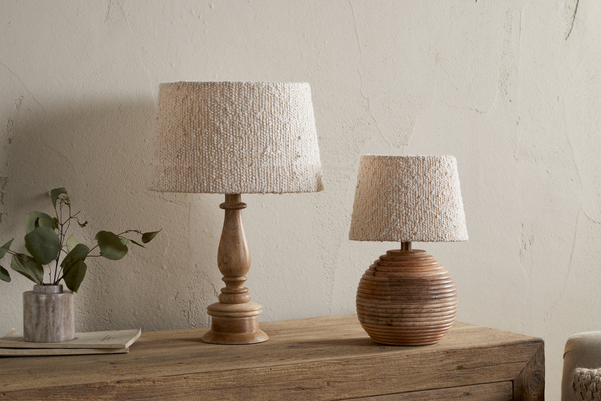 Two nkuku Sakiti Bouclé Lampshades in off white provide soft lighting on a rustic wooden table, each with a unique base. A small vase with greenery rests nearby, set against a textured off-white wall.