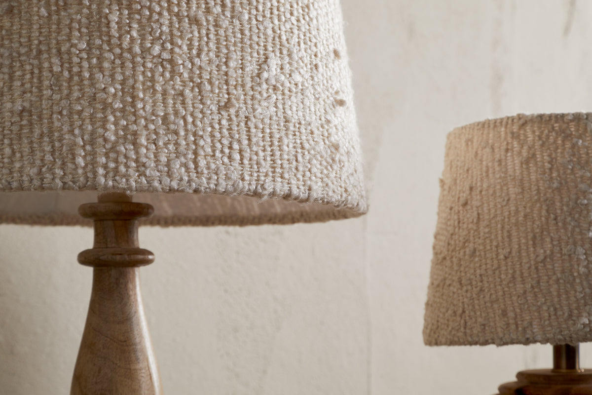 Close-up of two nkuku Sakiti Bouclé Lamps with off-white, textured woven lampshades and wooden bases. One lamp stands prominently in the foreground, while the other is partially visible against a neutral wall.