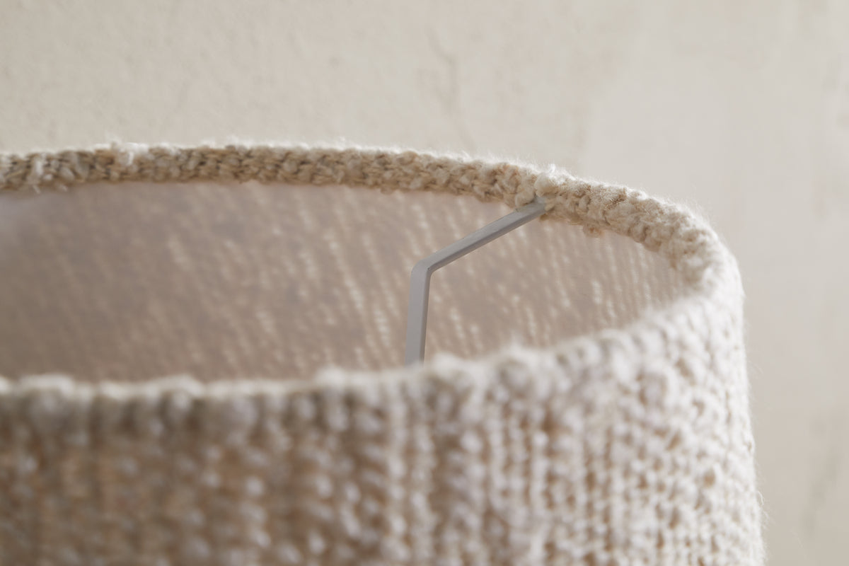 Close-up of the top edge of the Sakiti Bouclé Lampshade in off white by nkuku, featuring textured woven fabric. A metal support arm is visible inside, with soft ambient lighting casting gentle shadows on its surface and the wall.