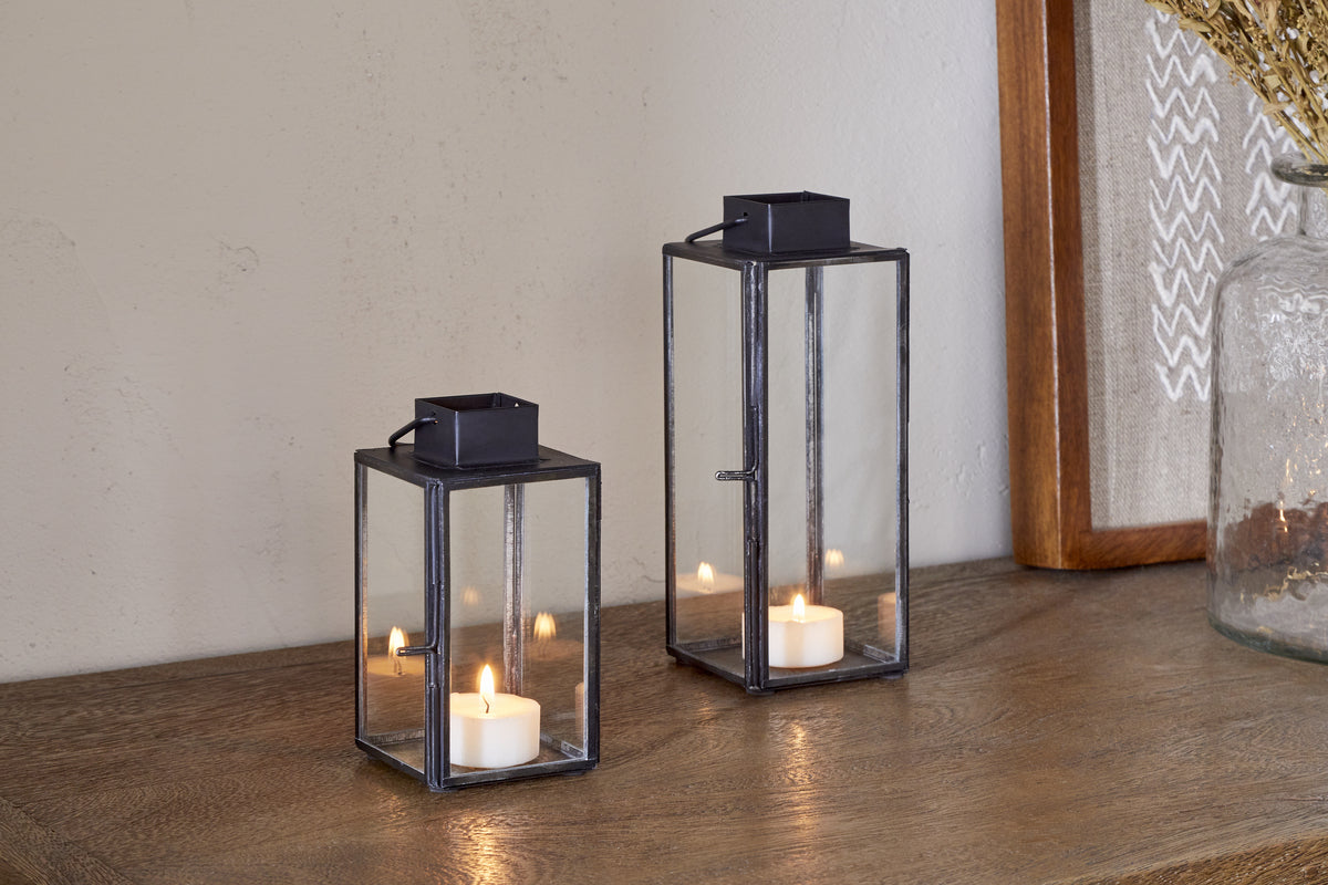 Two nkuku Sia Mini Table Lanterns in black, each holding a lit tealight, are arranged on a wooden surface beside a framed artwork and a glass jar of dried flowers.