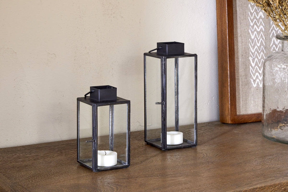 Two nkuku Sia Mini Table Lanterns in black glass and metal—one short, one tall—rest on a wooden surface, each holding a white tealight. Decorative items appear partially in the background.