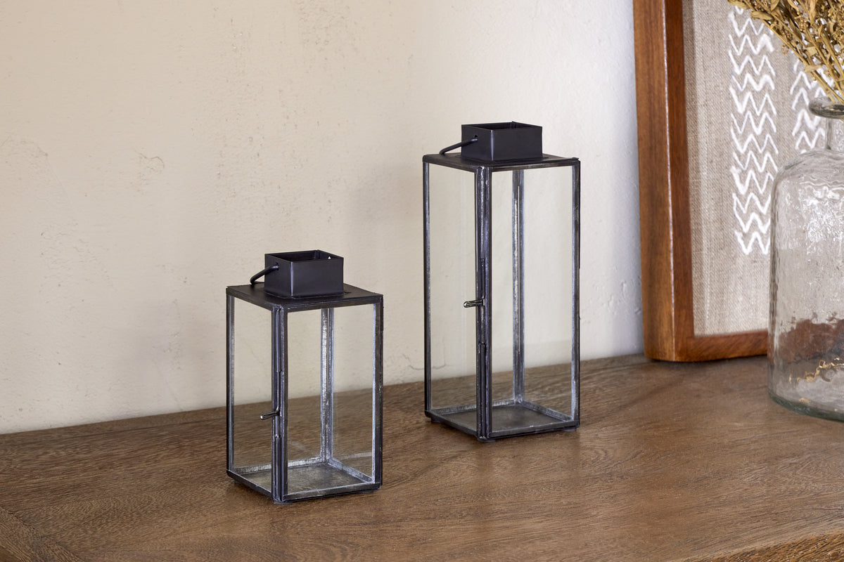 Two Sia Mini Table Lanterns by nkuku, featuring black metal frames, rest on a wooden surface near a glass vase and framed artwork—perfect for holding tealights and enhancing your decor.