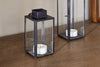 A Sia Mini Table Lantern by nkuku in antique black metal and glass sits on a wooden surface with a white tealight candle inside, next to a taller lantern of similar style.