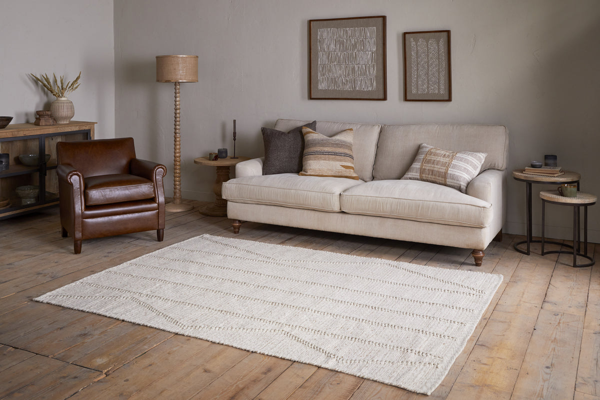 A cozy living room with a cream sofa, neutral pillows, a brown leather armchair, wooden side tables, and framed art on beige walls features the nkuku Shanti Wool & Cotton Rug - Natural on wood floors with warm, minimal decor.