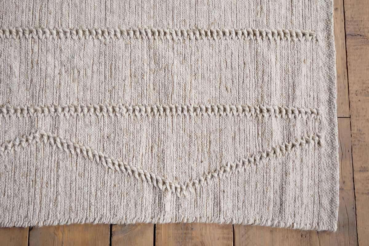 A close-up of the Shanti Wool & Cotton Rug - Natural by nkuku showcases its textured zigzag pattern and natural fiber look, set on a medium brown wooden floor.