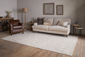 A cozy living room with a beige sofa, brown leather armchair, neutral pillows, wooden side tables, decorative wall art, wooden floors, and the nkuku Shanti Wool & Cotton Rug in Natural.