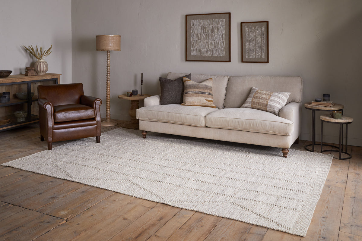 A cozy living room with a beige sofa, brown leather armchair, neutral pillows, wooden side tables, decorative wall art, wooden floors, and the nkuku Shanti Wool & Cotton Rug in Natural.