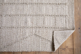 The Shanti Wool & Cotton Rug - Natural by nkuku, featuring subtle geometric patterns, is spread on a wooden floor with its bottom right corner folded to reveal the stylish underside.