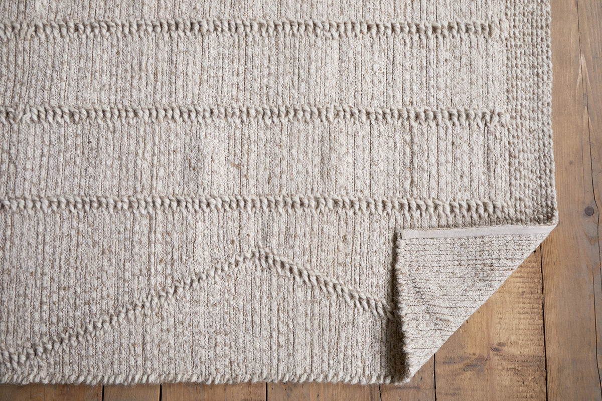 The Shanti Wool & Cotton Rug - Natural by nkuku, featuring subtle geometric patterns, is spread on a wooden floor with its bottom right corner folded to reveal the stylish underside.