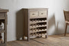 The Shanthi Reclaimed Wood Wine Rack - Natural by nkuku stands against a white wall on a wooden floor, featuring two small drawers and horizontal slots for wine storage. A woven chair and other furniture are partially visible nearby.