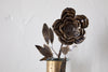The nkuku Svara Stem Rose in Antique Brass showcases layered metallic petals, a decorative stem with leaves, and is displayed in a small glass vase against a plain white background.