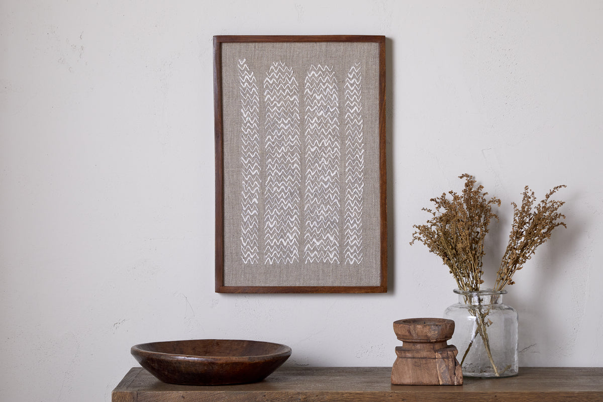 The Samaya Hand Stitched Wall Art - Natural by nkuku, featuring a white zigzag on beige, hangs above a wooden surface with a sustainable mango wood bowl, two stacked wooden pieces, and a glass jar of dried flowers.