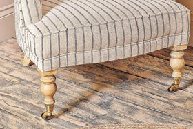 The nkuku Sanja Stripe Armchair, with its curved beige striped fabric and decorative wooden legs featuring brass front casters, sits on a wooden floor with a woven rug edge visible in the corner.