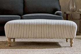 The nkuku Sanja Stripe Upholstered Ottoman in Off White, featuring a natural linen stripe design and four turned wooden legs, sits on a textured rug in front of a dark gray sofa.