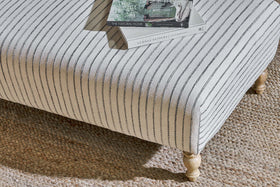 The nkuku Sanja Stripe Upholstered Ottoman in Off White features light wood legs and sits on a woven jute rug, topped with two books including “THE NATURAL HOME,” to showcase a cozy, natural linen-inspired minimalist style.