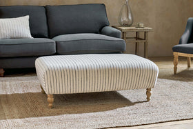 The Sanja Stripe Upholstered Ottoman by nkuku, in off white, features a subtle stripe design and short wooden legs. It sits on a woven rug before a dark gray sofa with a striped pillow, next to a wooden side table with a glass vase.