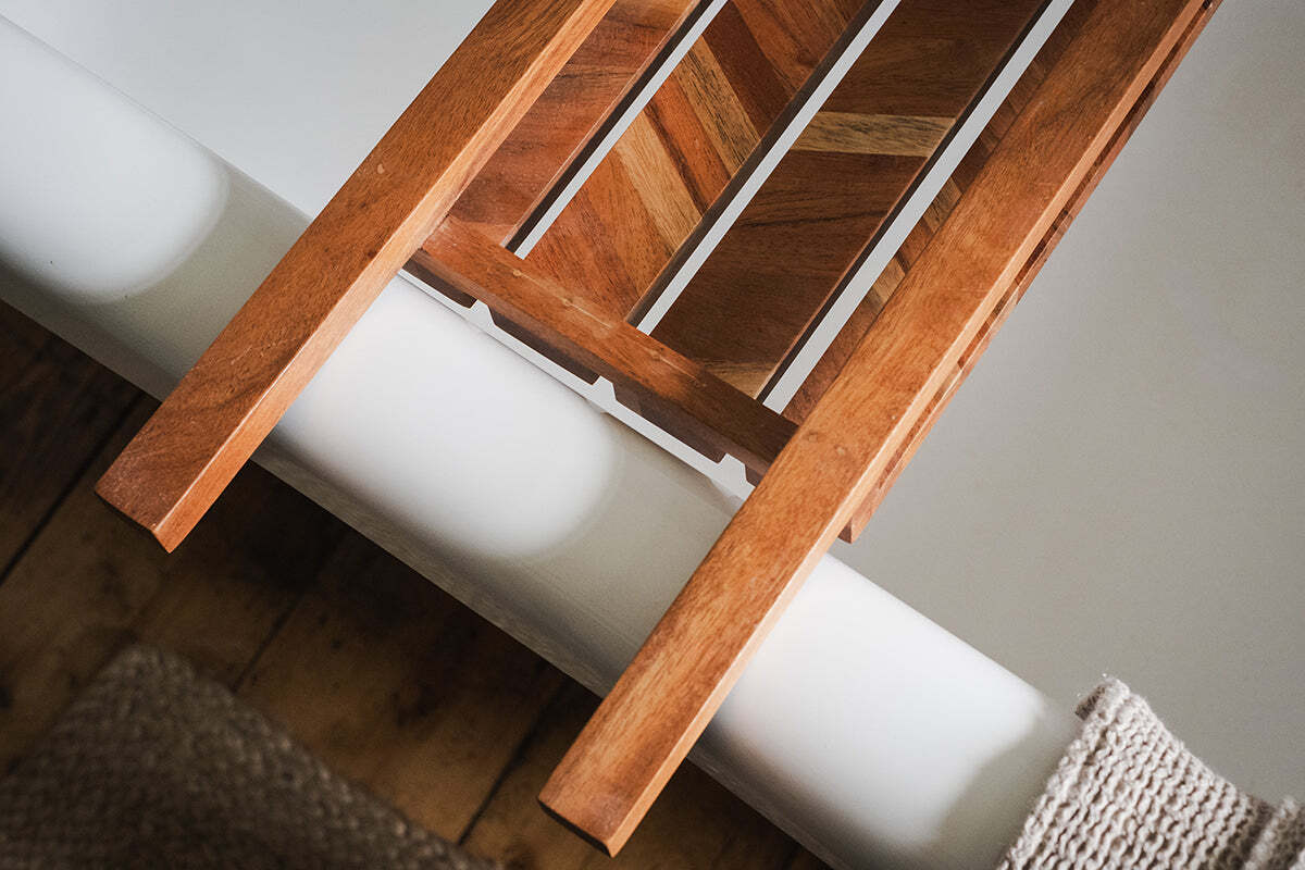 A close-up of the nkuku Sukha Mango Bath Bridge - Natural, featuring a slatted chevron design, rests on a white tub in a spa-like bathroom with a beige textured rug and wooden floor.