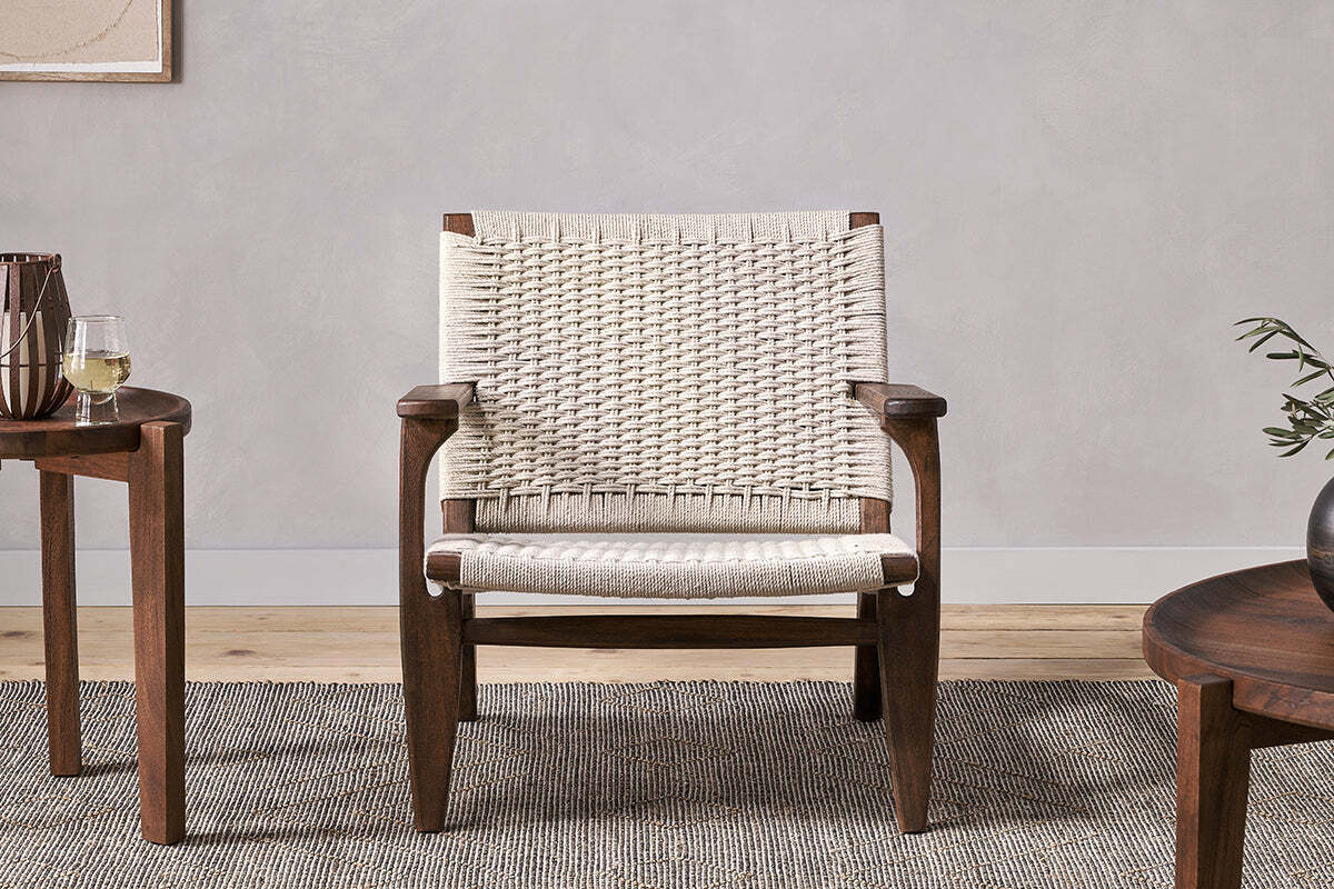 The Surat Woven Lounger - Natural by nkuku features acacia wood, wide armrests, and a hand-woven cotton rope seat and back. Placed on a textured gray rug, it sits between two round tables—one with white wine, the other with a vase of greenery.