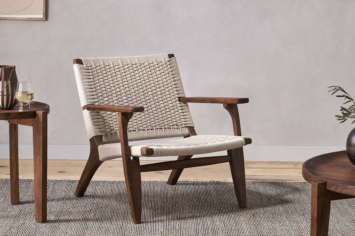 The Surat Woven Lounger - Natural by nkuku features acacia wood with a hand-woven cotton rope seat and backrest. Set on a gray rug, it’s styled with angled legs, armrests, and two wooden tables—one with a wine glass, the other with greenery.