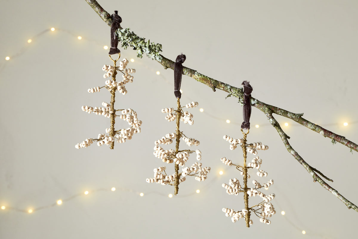 A mossy branch holds three nkuku Tanu Mango Wood Beaded Baubles in Brass & Natural, each shaped like a stylized tree with white beads, hanging from dark ribbons. Warm, soft string lights glow softly in the blurred background. Set of 3.