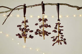 The nkuku Tanu Mango Wood Beaded Baubles - Brass & Ruby (Set of 3) feature ruby-toned and brass beads on gold wire, hanging from dark ribbons. Soft, warm lights glow in the background, adding a festive touch.