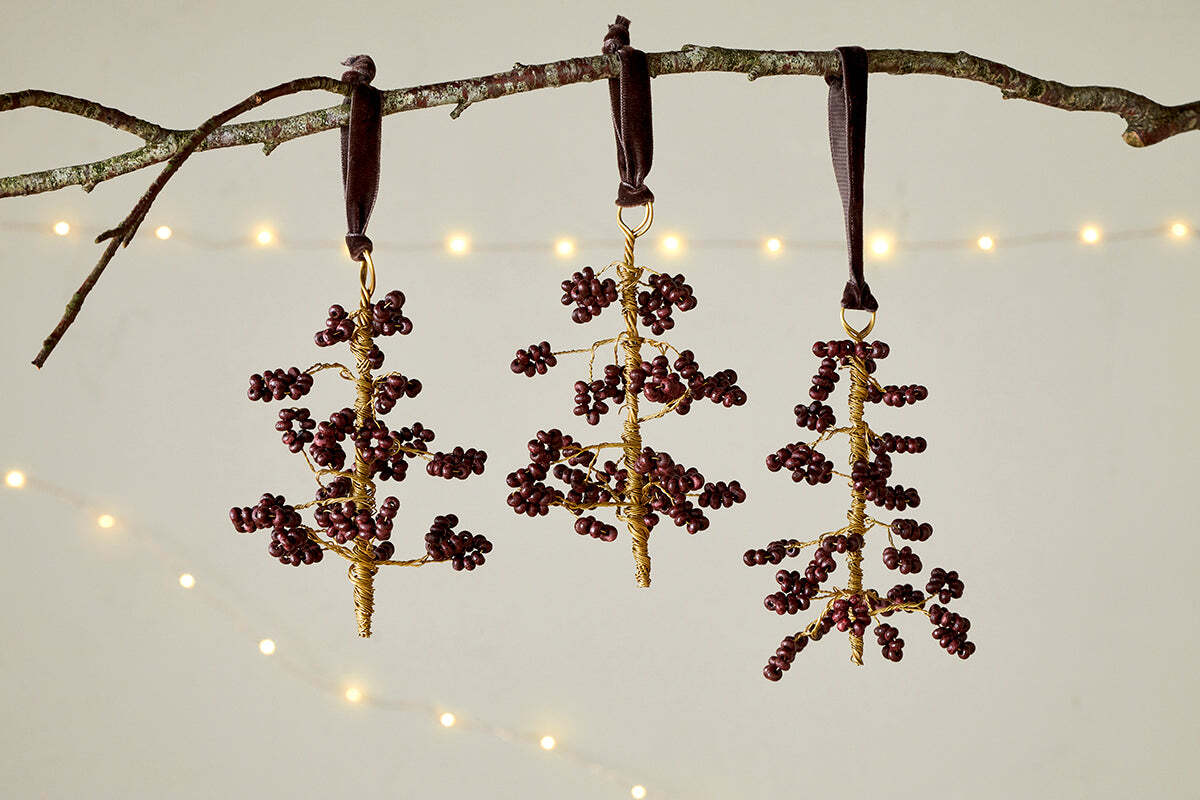 The nkuku Tanu Mango Wood Beaded Baubles - Brass & Ruby (Set of 3) feature ruby-toned and brass beads on gold wire, hanging from dark ribbons. Soft, warm lights glow in the background, adding a festive touch.