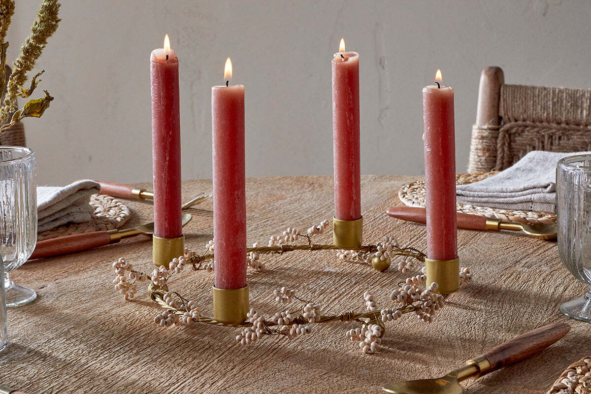 A rustic table features nkuku's Tanu Mango Wood Beaded Candle Holders in brass and natural, each with a lit red candle arranged in a circle, surrounded by a white flower garland, crystal glasses, napkins, and wooden-handled cutlery for a festive setting.