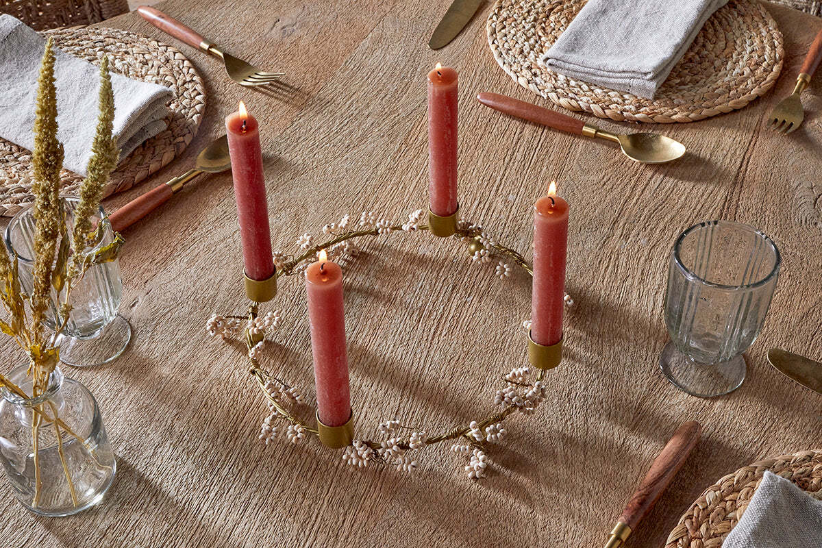 A mango wood table set for a meal features four nkuku Tanu Mango Wood Beaded Candle Holders in brass and natural, each topped with a red candle and surrounded by small white flowers. Place settings include woven placemats, gray napkins, gold utensils, and clear glasses.