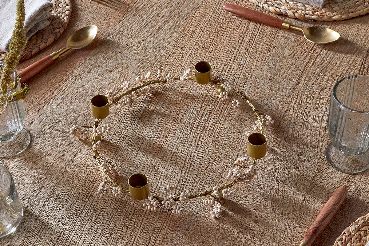 The nkuku Tanu Mango Wood Beaded Candle Holder in Brass & Natural sits on a rustic wooden table with woven placemats, mango wood and gold cutlery, clear glasses, and a beige napkin—ideal for softly lit festive tablescapes.