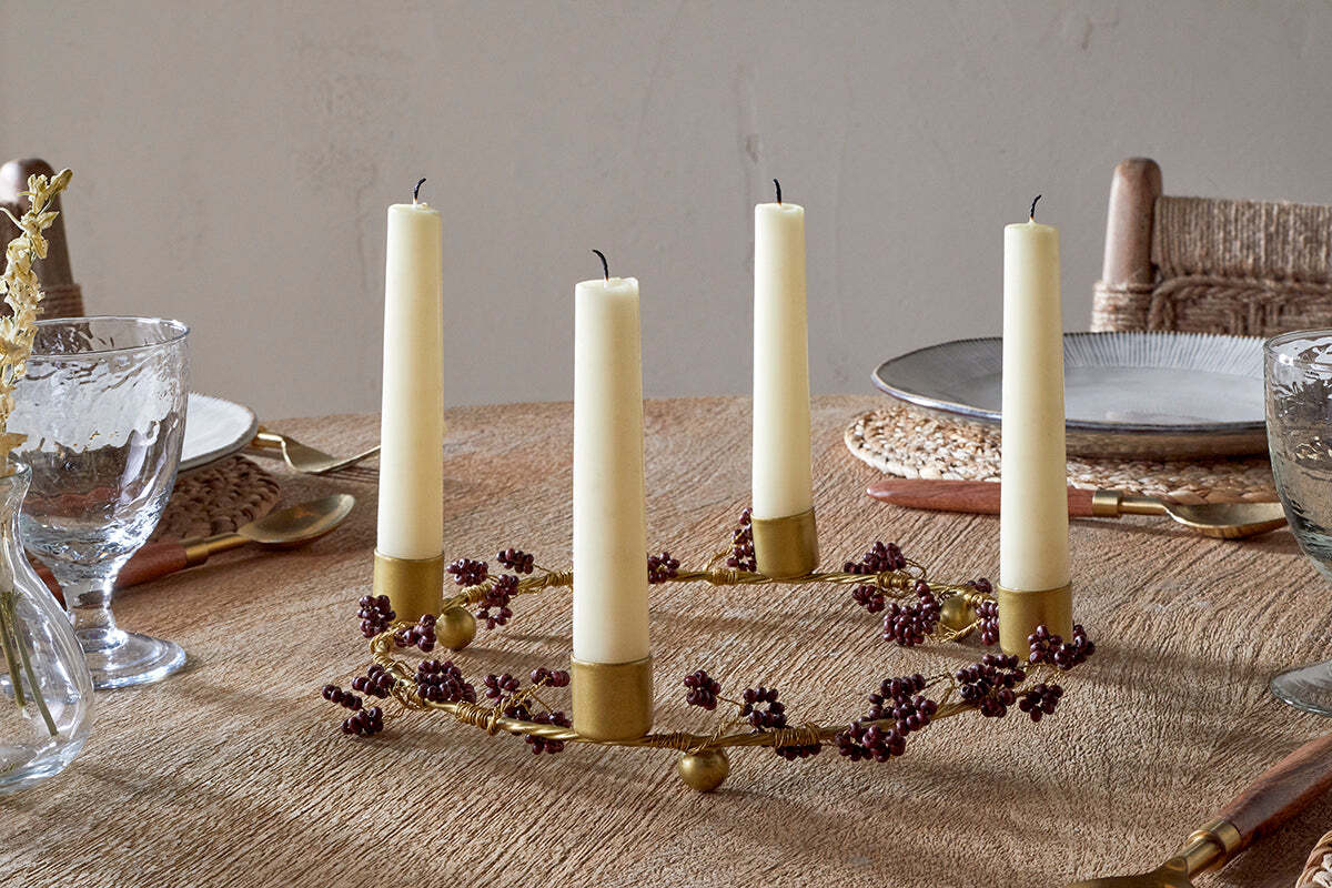 The nkuku Tanu Mango Wood Beaded Candle Holder in Brass & Ruby, adorned with small purple berries and cream candles, adds festive charm to a wooden dining table set with woven placemats, clear glasses, plates, and cutlery.
