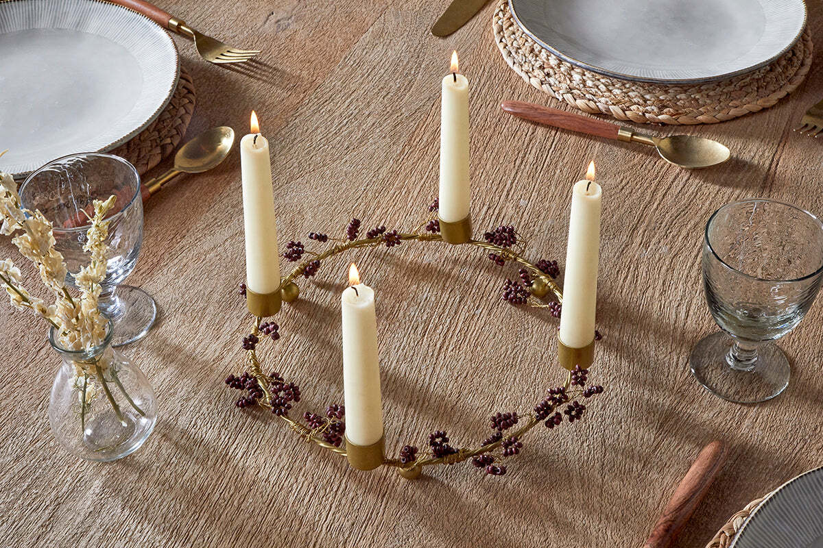 A round wooden table is set for dining, featuring the nkuku Tanu Mango Wood Beaded Candle Holder in Brass & Ruby with four cream candles and burgundy berries as a festive centerpiece. Nearby, dried flowers sit in a small vase.