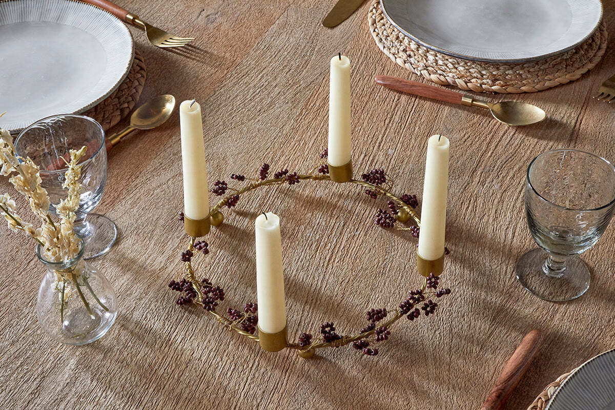 The nkuku Tanu Mango Wood Beaded Candle Holder in Brass & Ruby, styled with five cream candles and purple florals, is a stunning centerpiece for festive tablescapes. A small vase of dried flowers sits beside this elegant decorative accent.