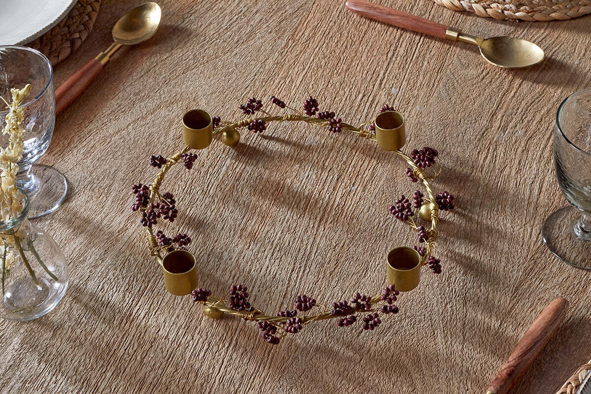 The Tanu Mango Wood Beaded Candle Holder in Brass & Ruby by nkuku adds a festive, rustic accent to a textured wooden table, surrounded by glassware and gold cutlery for a charming seasonal tablescape.