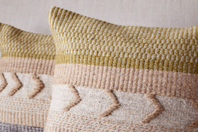 The nkuku Tuleeka Wool Cushion Cover in ochre features a textured woven design in beige, tan, and olive green with chevron-style raised details across the lower half. One decorative cover is shown prominently in the front.
