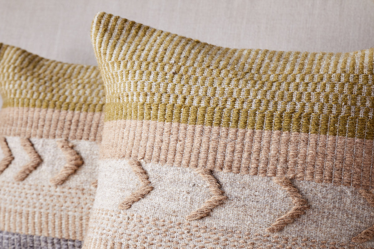 The nkuku Tuleeka Wool Cushion Cover in ochre features a textured woven design in beige, tan, and olive green with chevron-style raised details across the lower half. One decorative cover is shown prominently in the front.
